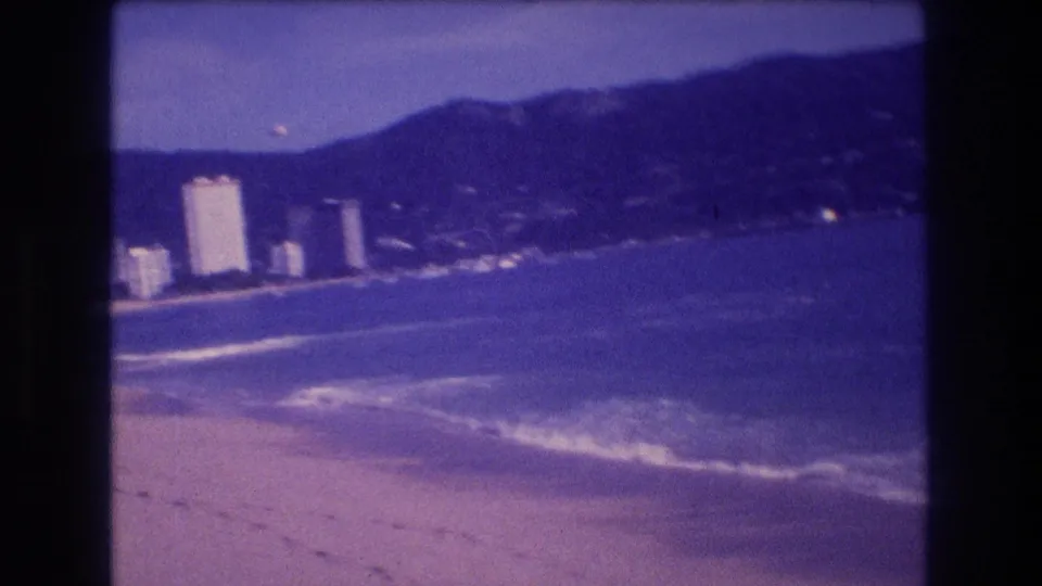 8mm home-movie frame showing a wider angle of Acapulco's bay, hotel cluster, and surrounding mountains under the characteristic aged-film blue-violet cast, circa 1982.