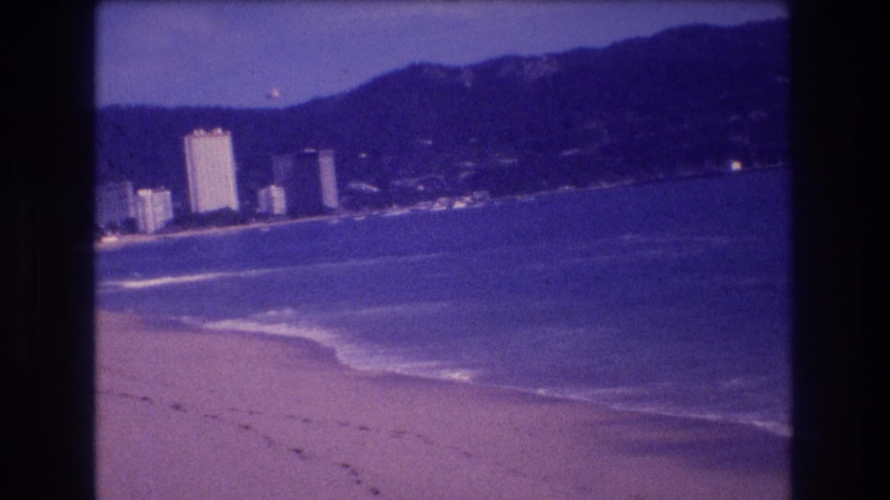 Closing 8mm home-movie frame of the Acapulco beach sequence circa 1982; hotel buildings and mountains under blue-violet aged-film cast with visible grain.