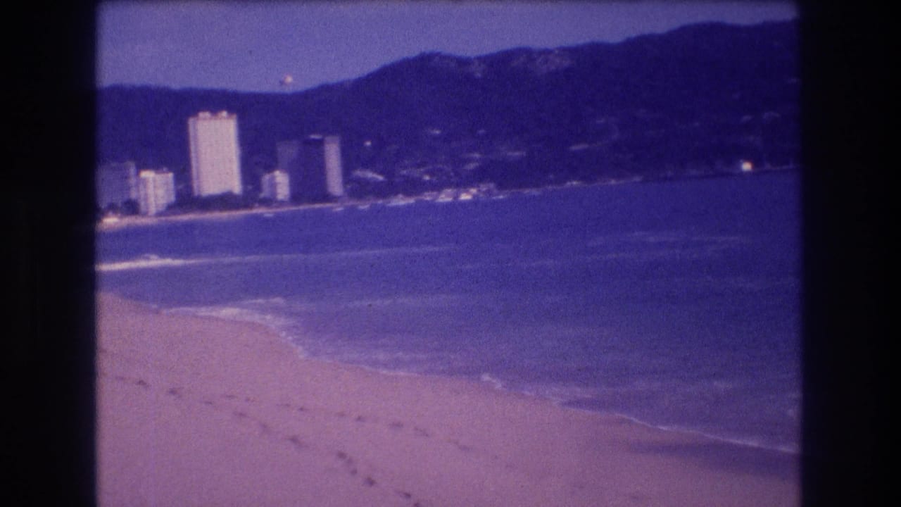 Sandy beach with footprints, gentle surf break, and coastal high-rise buildings beneath a dark mountain range, captured on 8mm film.