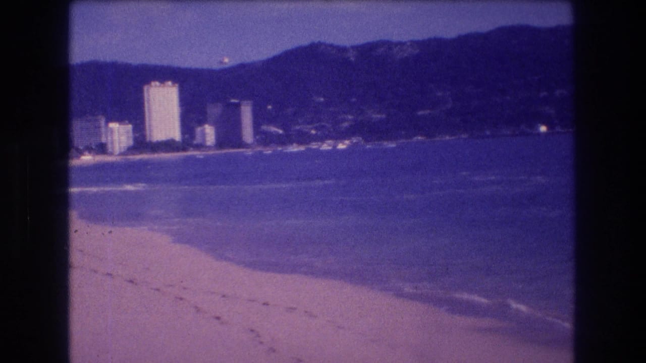 Sandy beach, gentle wave break, and a cluster of coastal high-rise buildings beneath a dark mountain range, 8mm with purple color cast.