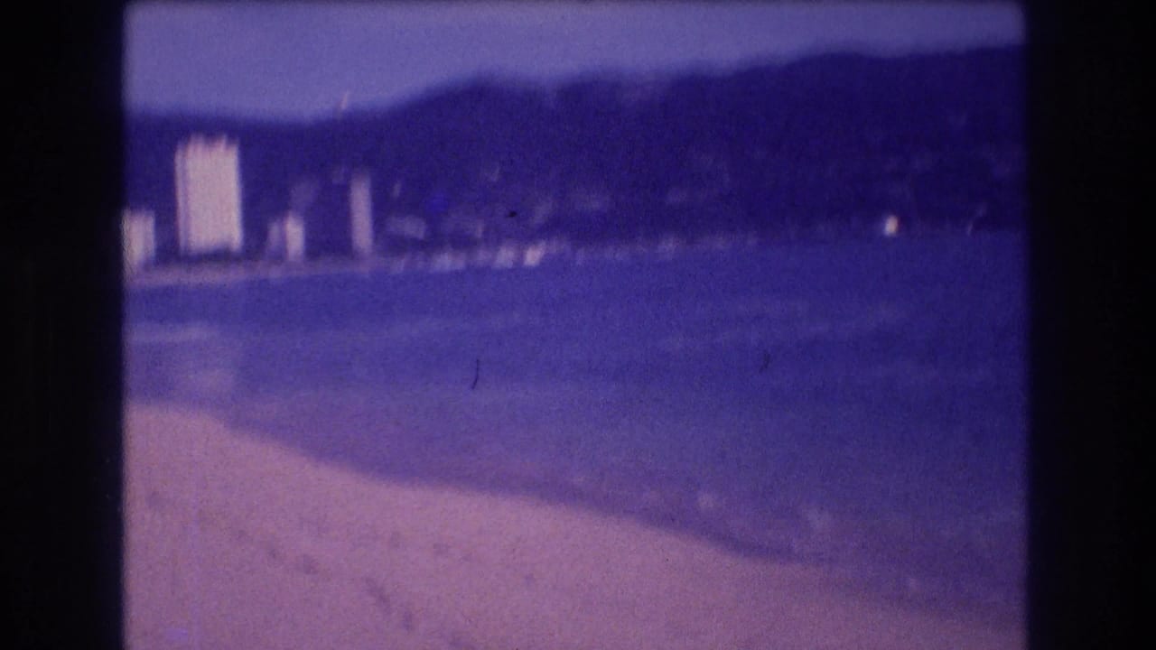 Motion-blurred 8mm frame from Acapulco beach, circa 1982, showing sandy shore, indistinct resort buildings, and mountain backdrop under a blue-purple haze.