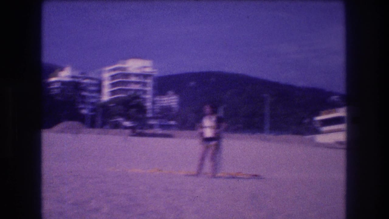 A woman stands on a sandy beach in light top and dark shorts, with resort hotel towers, trees, and mountains visible behind her, circa 1982 Acapulco, captured on Super 8 film.