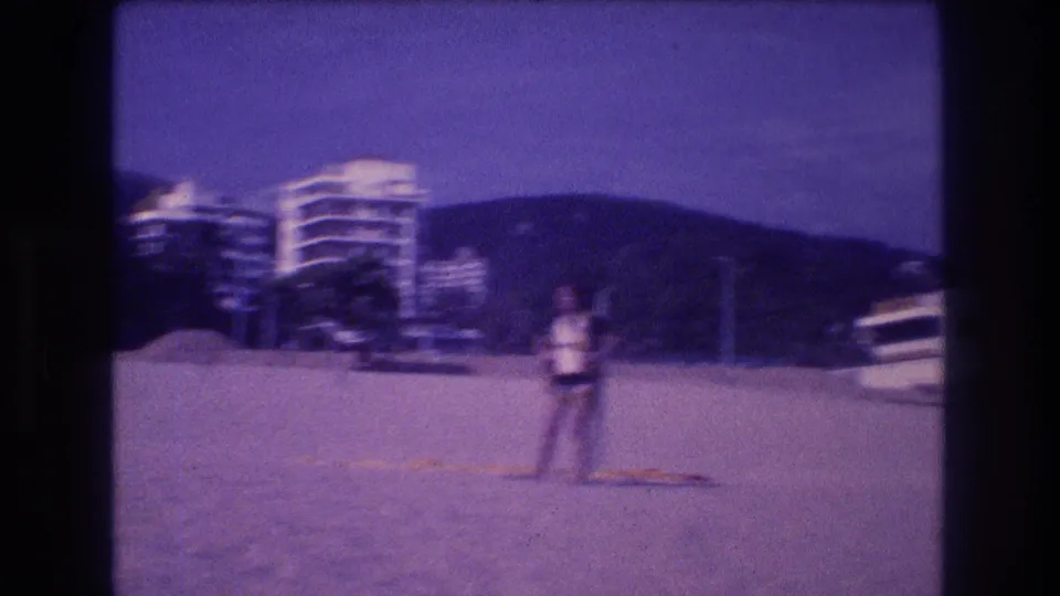 A woman stands on a sandy beach in light top and dark shorts, with resort hotel towers, trees, and mountains visible behind her, circa 1982 Acapulco, captured on Super 8 film.