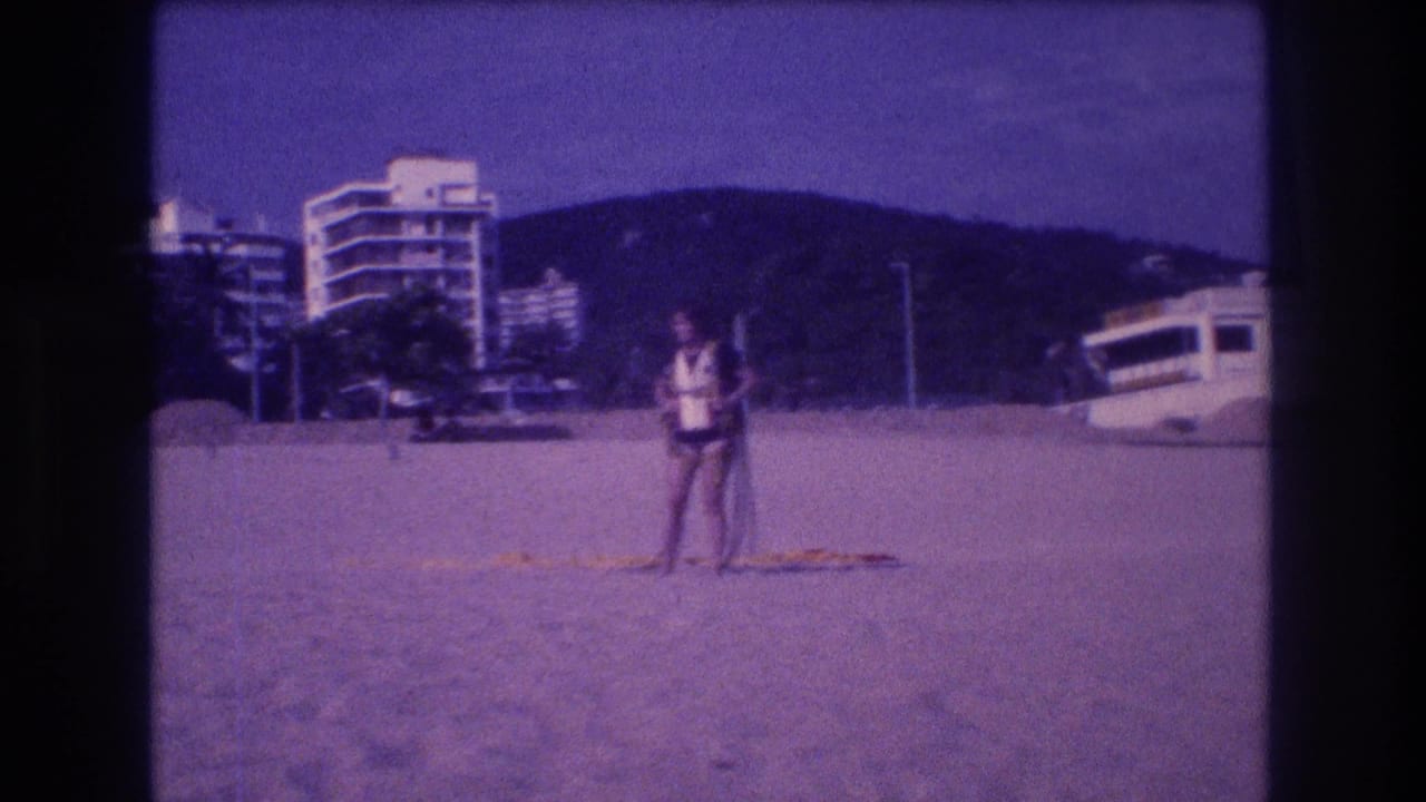 Adult figure on Acapulco beach, circa 1982, holding a long pole; multi-story resort hotels and coastal hills behind; aged 8mm purple-blue color cast.