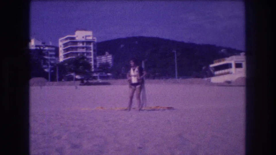 Adult figure on Acapulco beach, circa 1982, holding a long pole; multi-story resort hotels and coastal hills behind; aged 8mm purple-blue color cast.