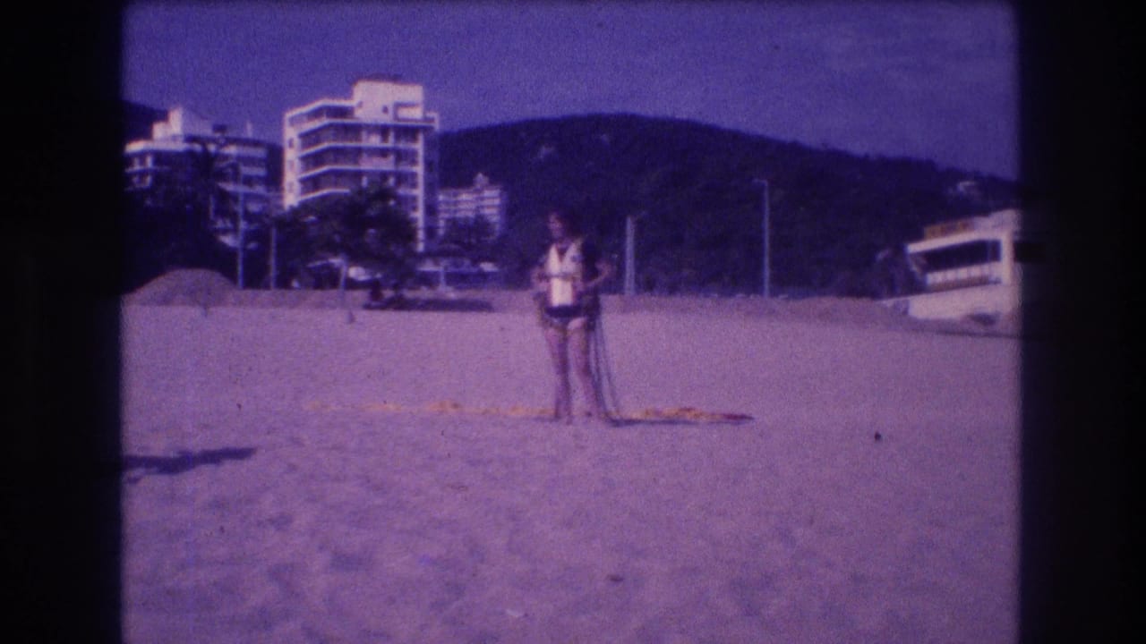 Adult figure on Acapulco beach, circa 1982, standing with long pole on open sand; balconied hotel skyline and coastal hills visible.