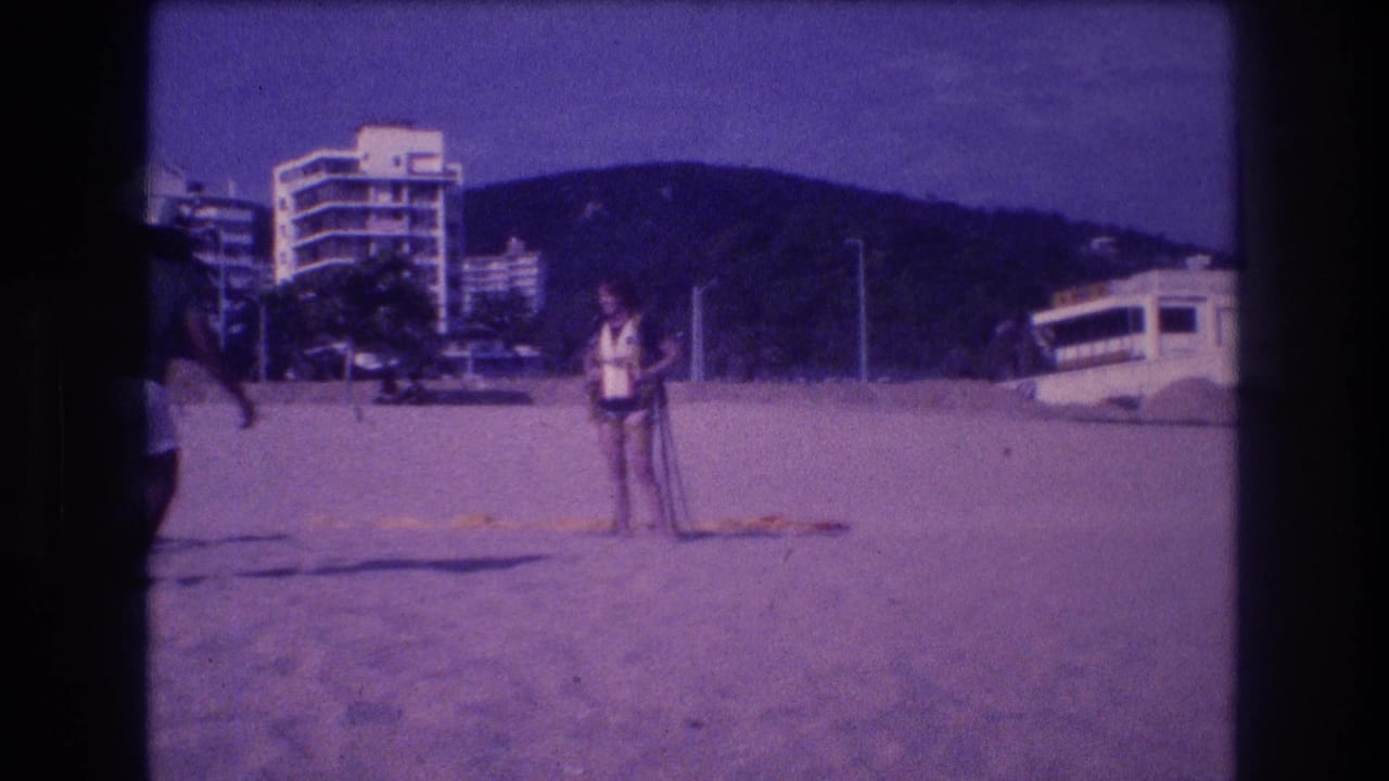 Adult figure on Acapulco beach circa 1982; dark circular film artifact at lower-left frame edge; multi-story hotels and green hills behind.