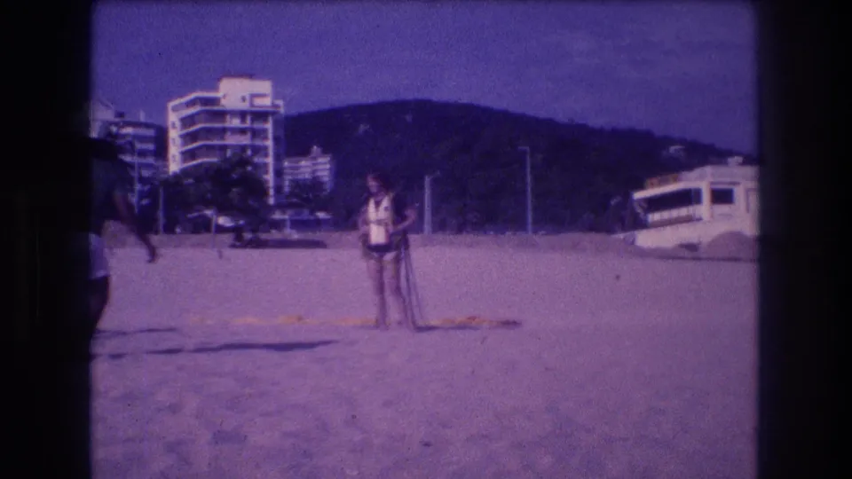 Adult figure on Acapulco beach circa 1982; dark circular film artifact at lower-left frame edge; multi-story hotels and green hills behind.