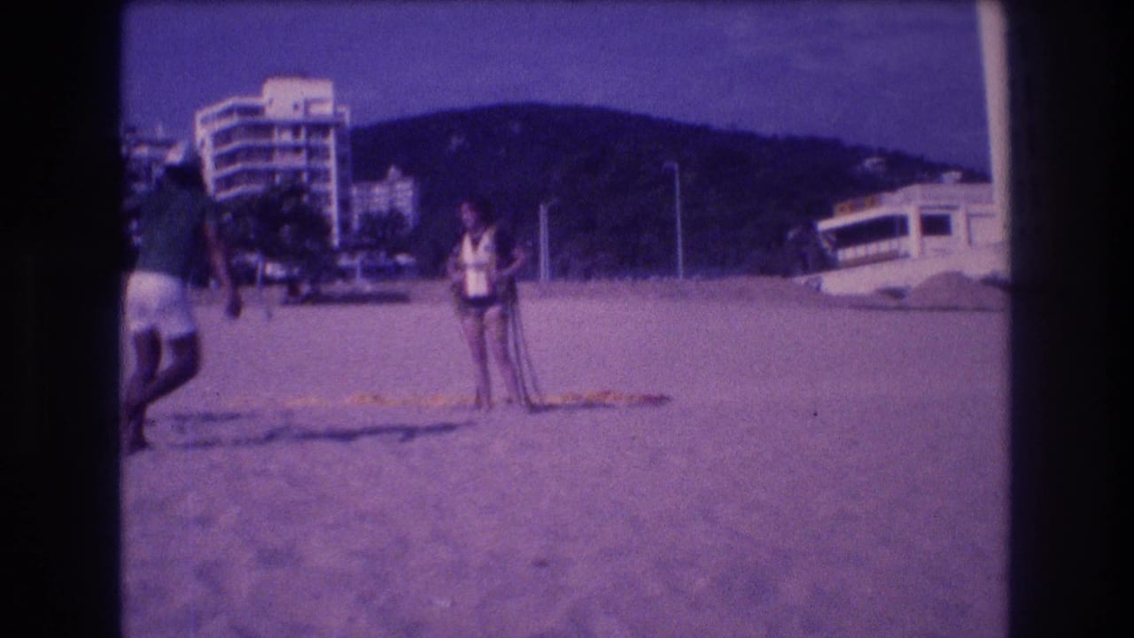 Two adults on a sandy Acapulco beach in 1982: a man in green shirt and white shorts, a woman in a white life jacket, with high-rise resort towers behind them.