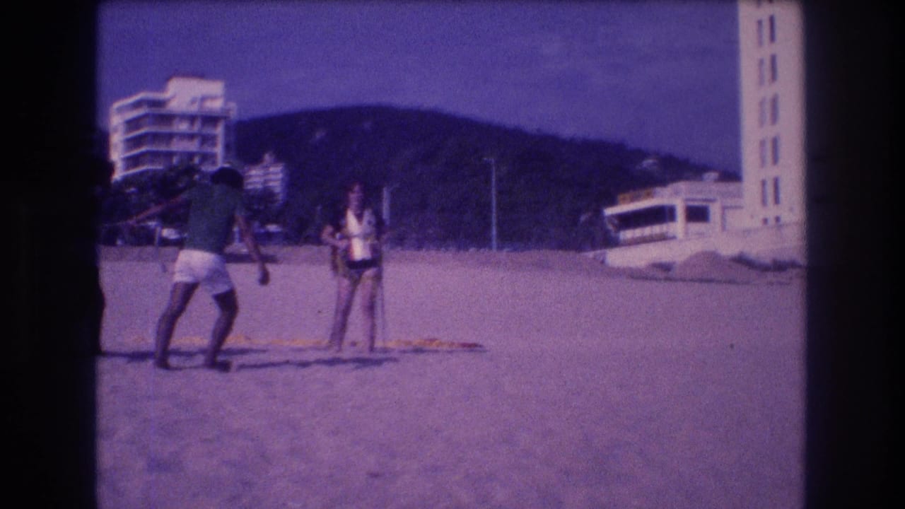 Continued shot of two adults on the Acapulco beach in 1982; the man adjusts his stance as the woman in a life jacket stands nearby, hotel towers and a green hill visible behind.