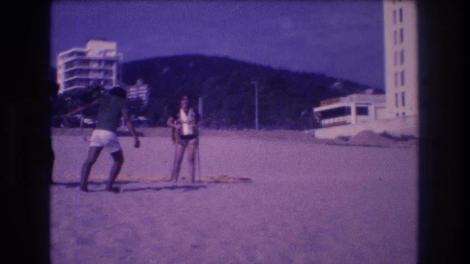 Continued shot of two adults on the Acapulco beach in 1982; the man adjusts his stance as the woman in a life jacket stands nearby, hotel towers and a green hill visible behind.