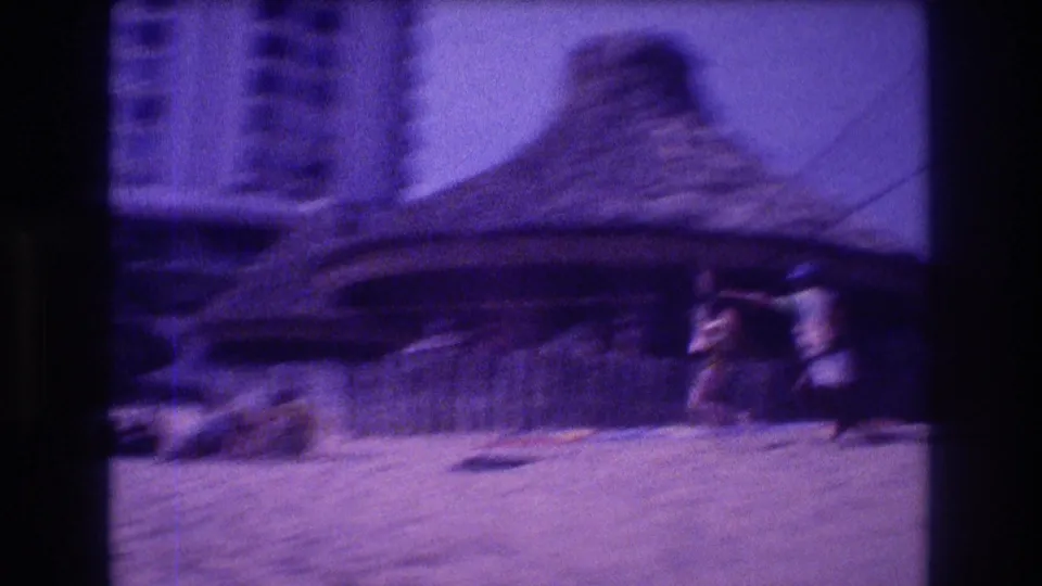 Motion-blurred 8mm frame panning past a large thatched palapa hut in Acapulco, 1982, with a person moving rapidly through frame suggesting the zip-line ride in progress.