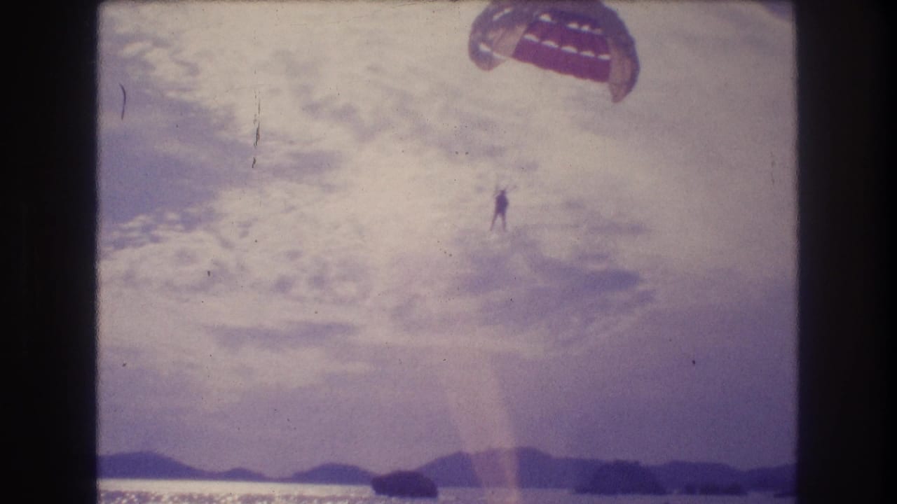 8mm home-movie frame circa 1982 showing a parasailer with reddish-orange dome canopy above a hill-lined coastline, consistent with Acapulco, Mexico; strong lens flare present.