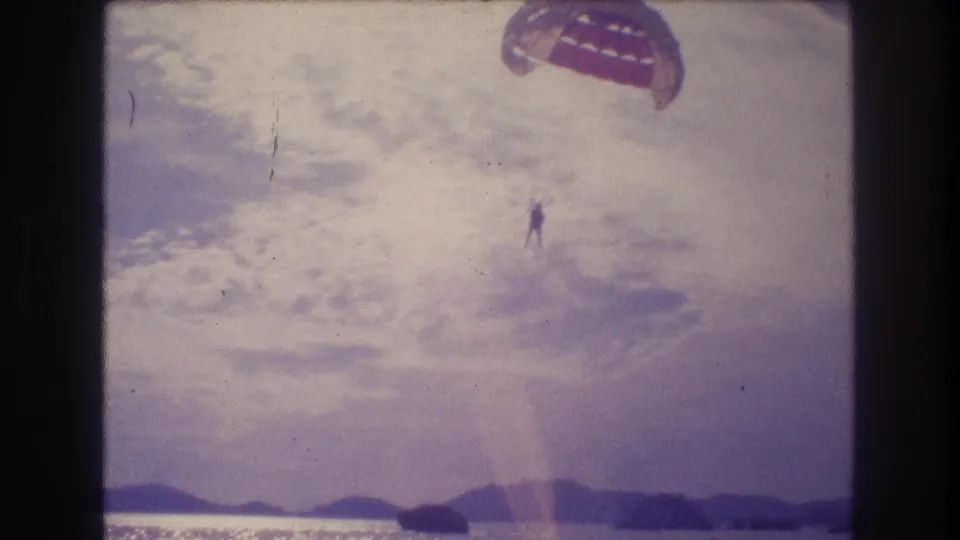 8mm home-movie frame circa 1982 showing a parasailer with reddish-orange dome canopy above a hill-lined coastline, consistent with Acapulco, Mexico; strong lens flare present.