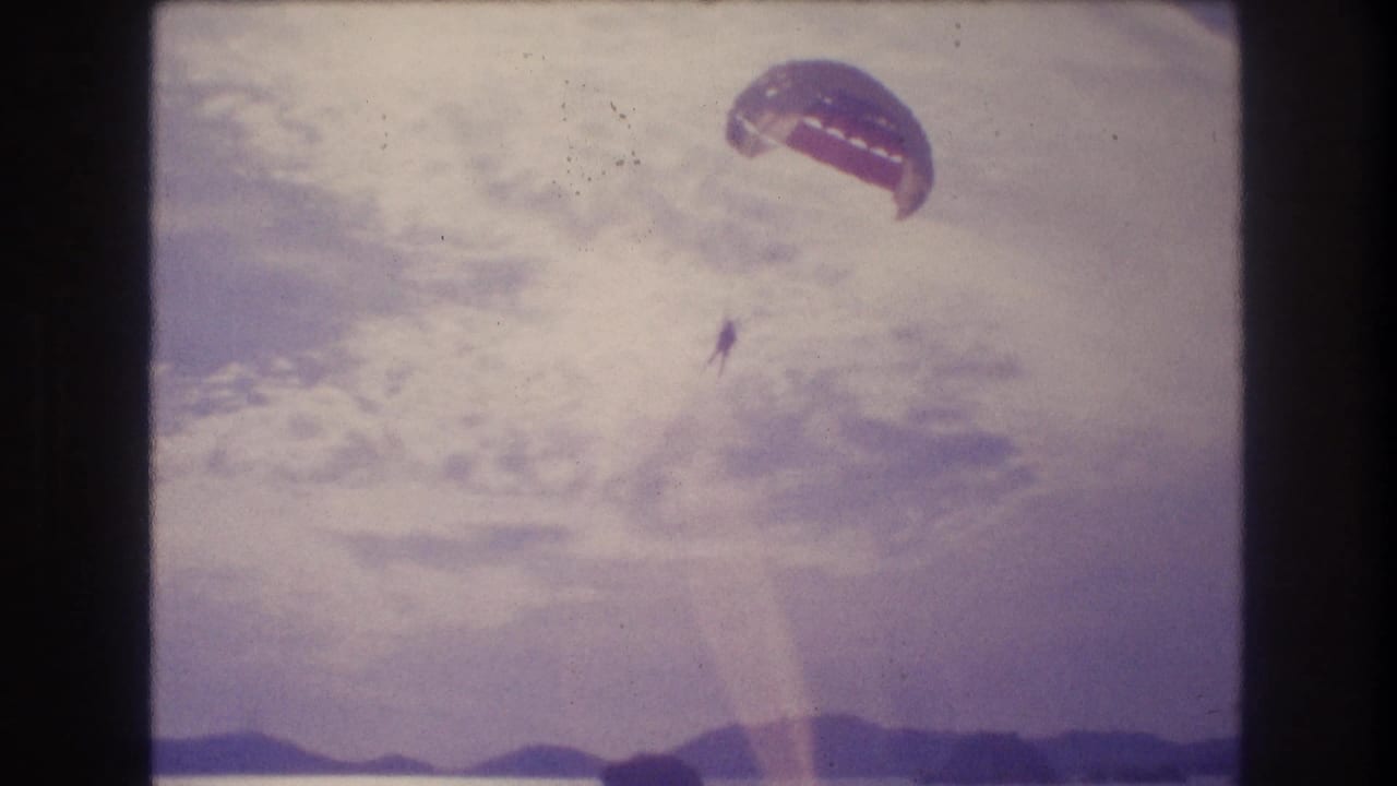 8mm home-movie frame circa 1982 showing a parasailer with dome canopy above a coastal hillline; diagonal lens flare crosses the frame; location consistent with Acapulco, Mexico.