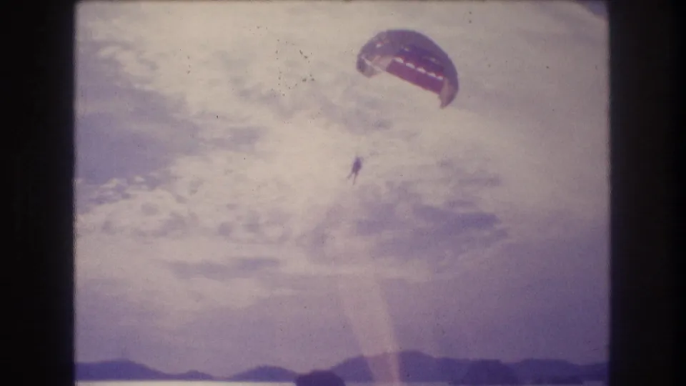 8mm home-movie frame circa 1982 showing a parasailer with dome canopy above a coastal hillline; diagonal lens flare crosses the frame; location consistent with Acapulco, Mexico.