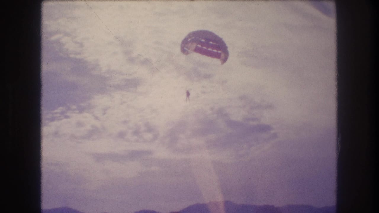 Mid-descent 8mm frame circa 1982 showing a parasailer with reddish dome canopy against a heavily overexposed sky; coastline no longer visible; consistent with Acapulco area.