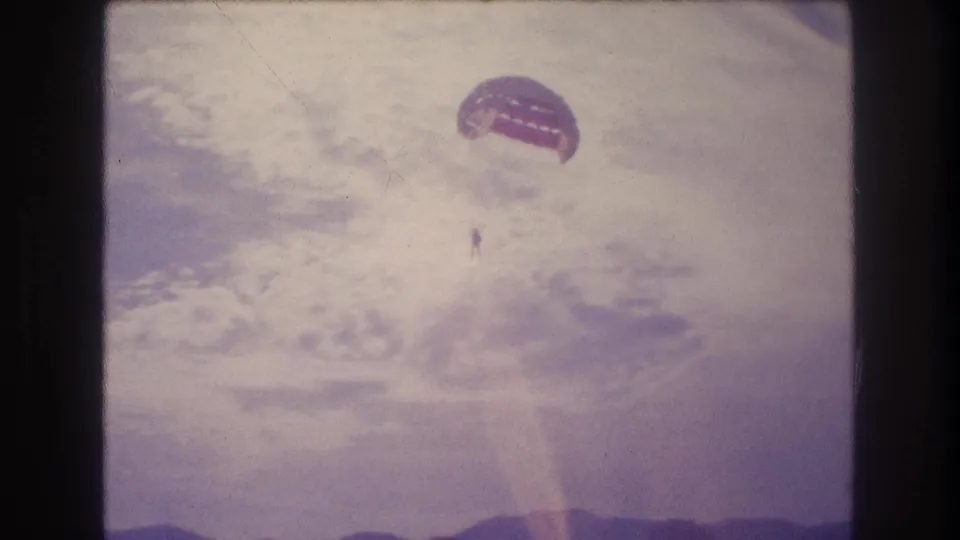Mid-descent 8mm frame circa 1982 showing a parasailer with reddish dome canopy against a heavily overexposed sky; coastline no longer visible; consistent with Acapulco area.
