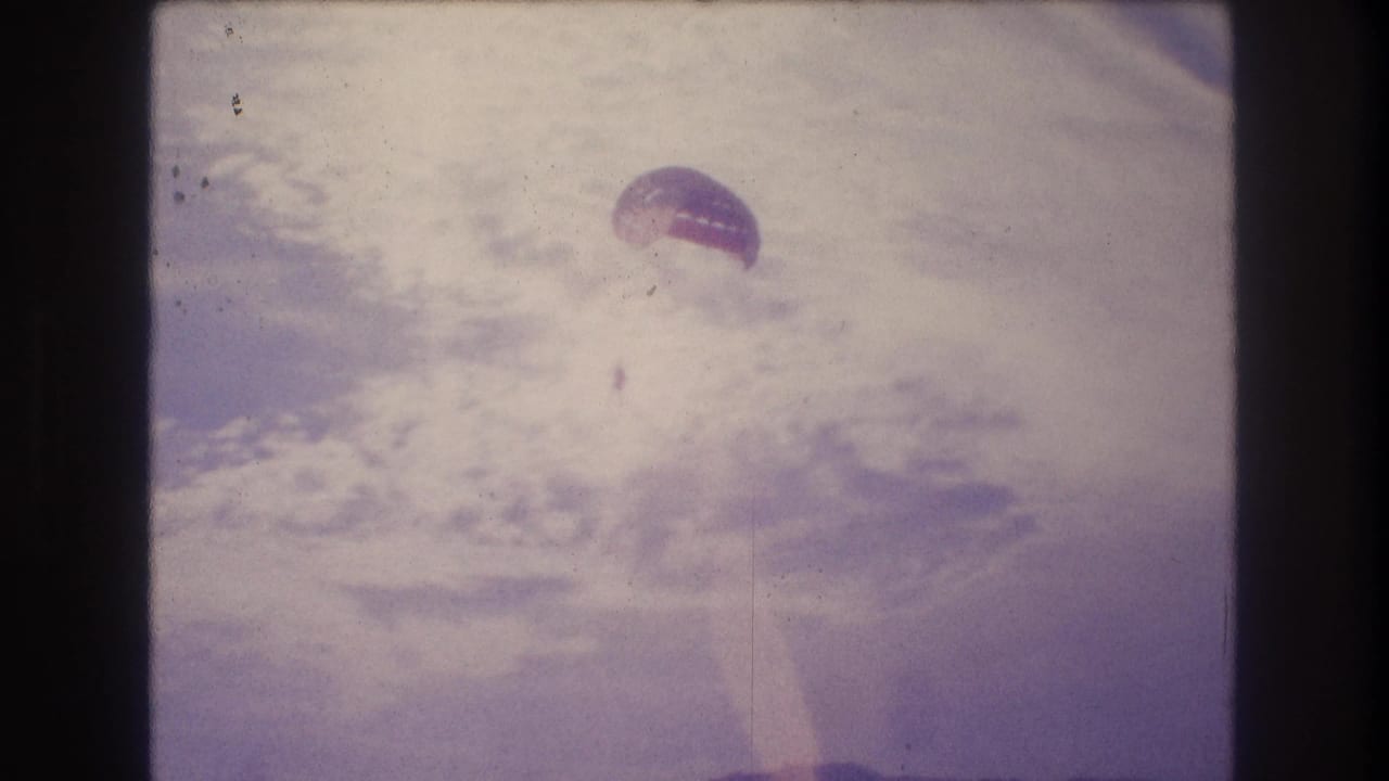 Final 8mm frame circa 1982 showing a parasailer with dome canopy against an overexposed washed-out sky; film dust and strong color fading consistent with 1980s consumer film stock.