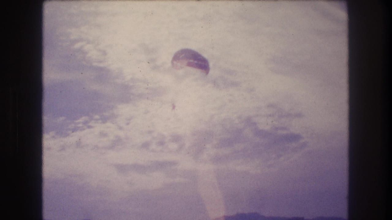 8mm home-movie frame from approximately 1982 showing a parachute canopy against a heavily bleached sky and coastal hillscape, consistent with Acapulco, Mexico.