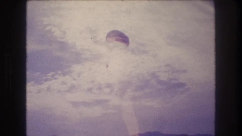 8mm home-movie frame from approximately 1982 showing a parachute canopy against a heavily bleached sky and coastal hillscape, consistent with Acapulco, Mexico.