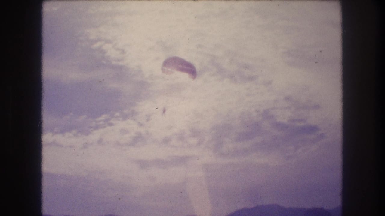 8mm home-movie frame circa 1982 showing a parachute drifting across a color-bleached sky over a coastal bay, consistent with Acapulco, Mexico.