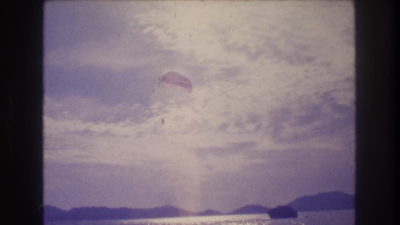 8mm home-movie frame circa 1982 showing a parachute high over a coastal bay with silhouetted hills and a possible shoreline structure at the lower right, consistent with Acapulco, Mexico.