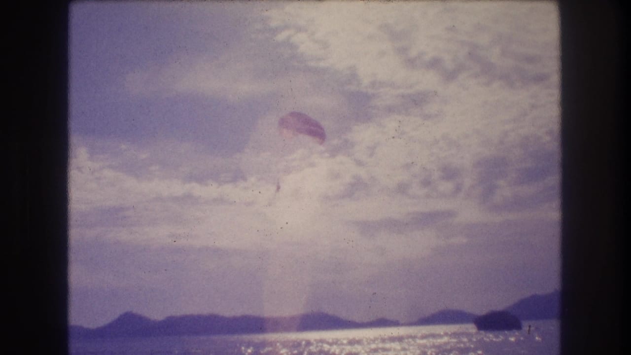 Small parachute canopy with figure suspended over sparkling ocean, overcast sky, distant mountain ridge, purple-blue 8mm color cast.