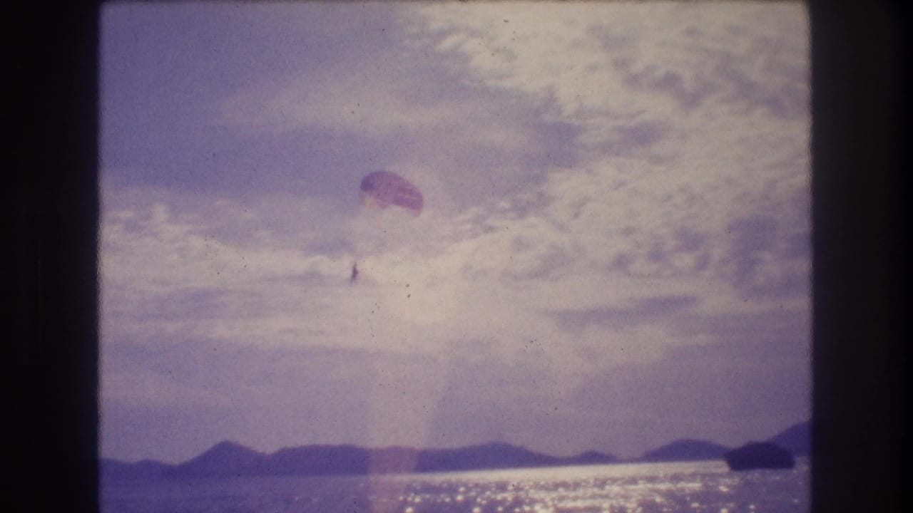 Parachute canopy with figure drifting upper-right against cloudy sky above sunlit ocean, mountain silhouette on horizon, 8mm color shift.