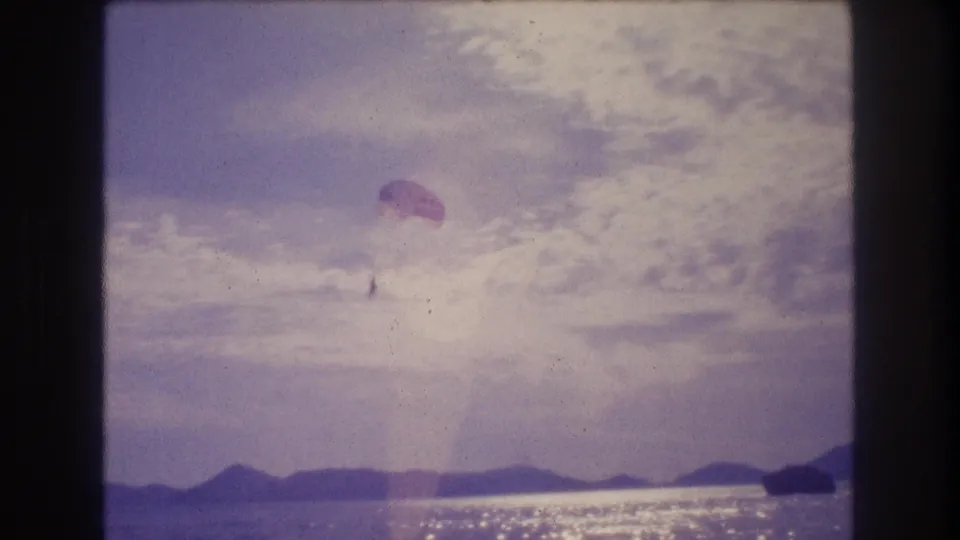 8mm home movie, Acapulco 1982 — parasail canopy in upper-right of frame against cloud-filled sky, sun-sparkling Pacific below and mountain ridge at horizon.
