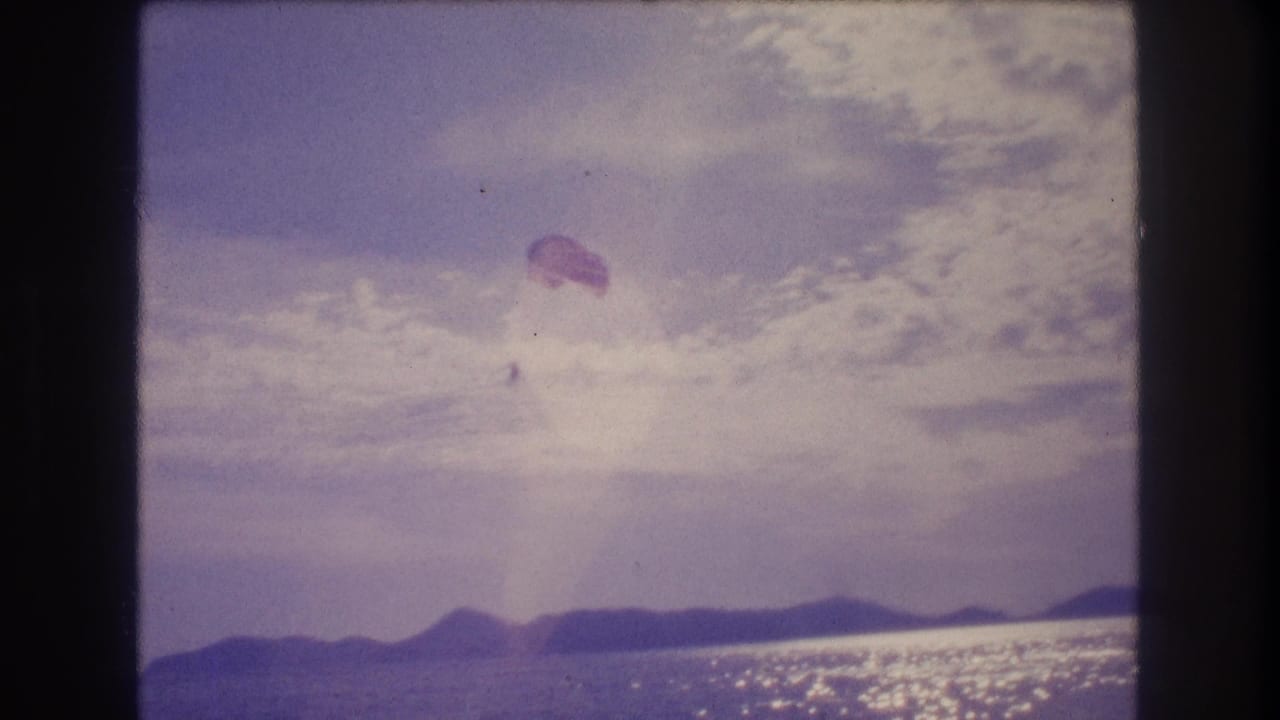 8mm home movie, Acapulco 1982 — parasail canopy now lower in the frame, appearing to descend toward the ocean surface, mountain ridge on horizon.