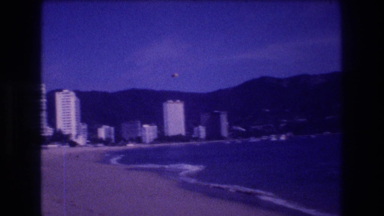 8mm home movie, Acapulco 1982 — panoramic beach view showing the hotel-zone high-rise strip against mountain hills under a vivid blue sky.