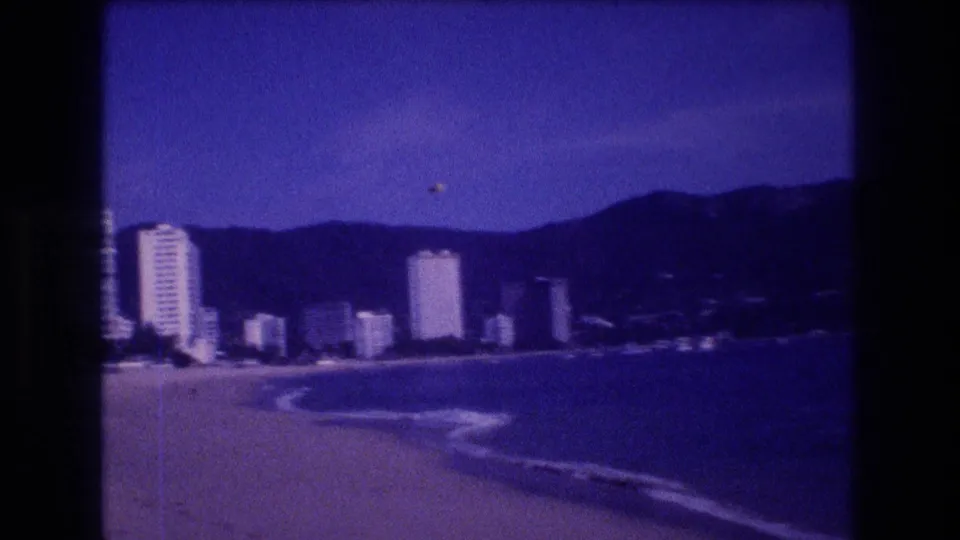 8mm home movie, Acapulco 1982 — panoramic beach view showing the hotel-zone high-rise strip against mountain hills under a vivid blue sky.