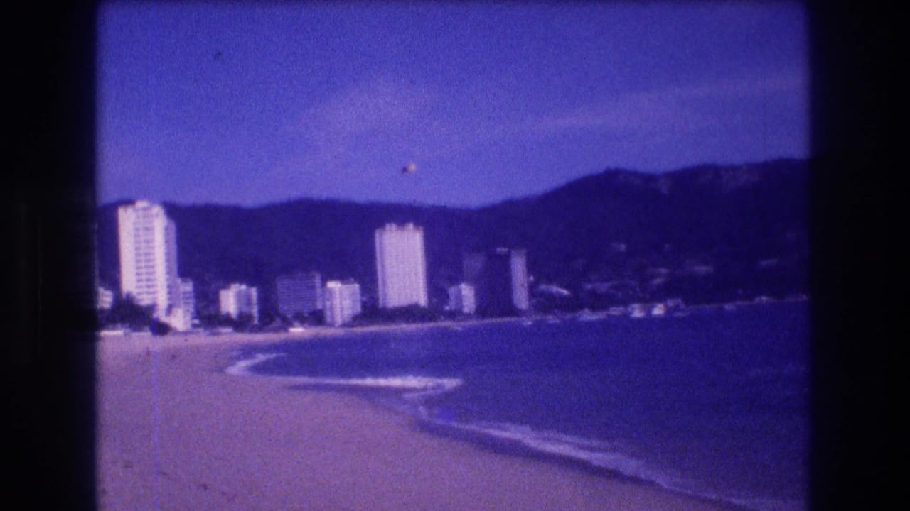 Sandy beach curving toward white high-rise hotels with forested mountains behind, heavy purple color cast and 8mm film grain.