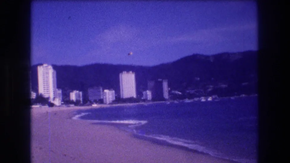 8mm home movie frame of Acapulco beach and hotel skyline, 1982, with characteristic purple dye-fade color cast and visible grain.