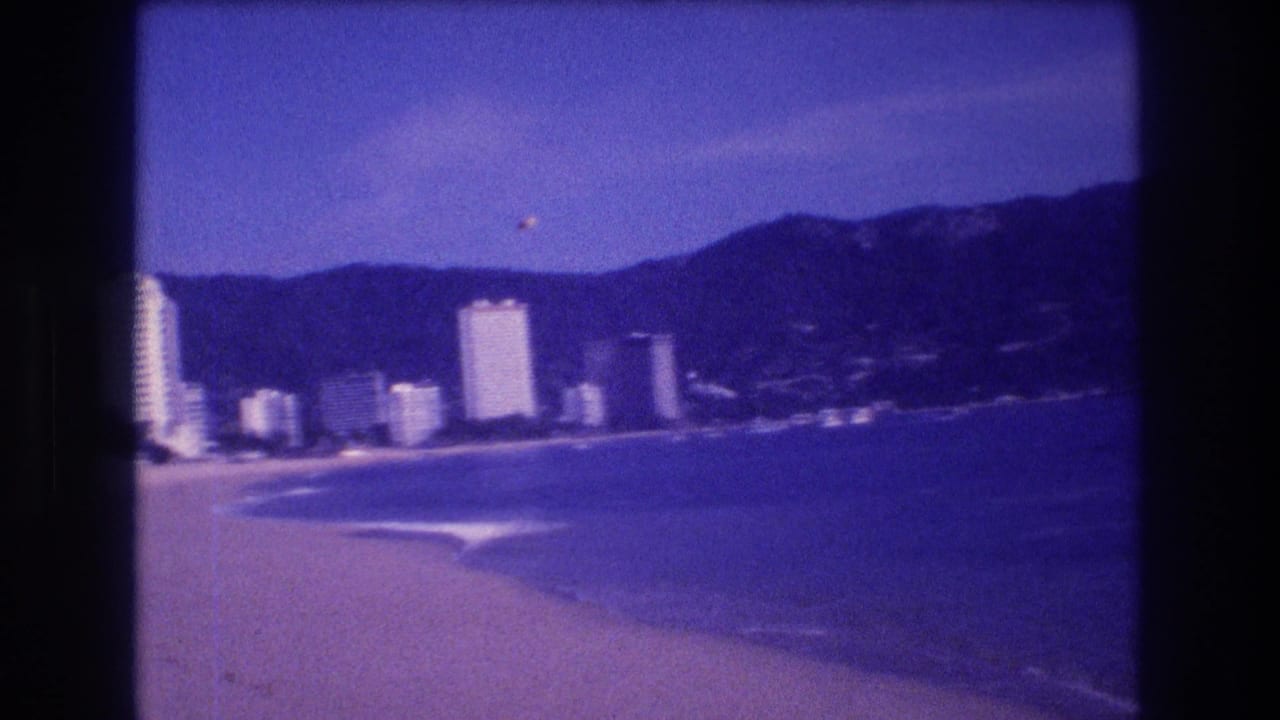 Beach panorama with hotel towers and mountains slightly right of previous frame, two small floating objects in upper sky, purple-toned 8mm stock.