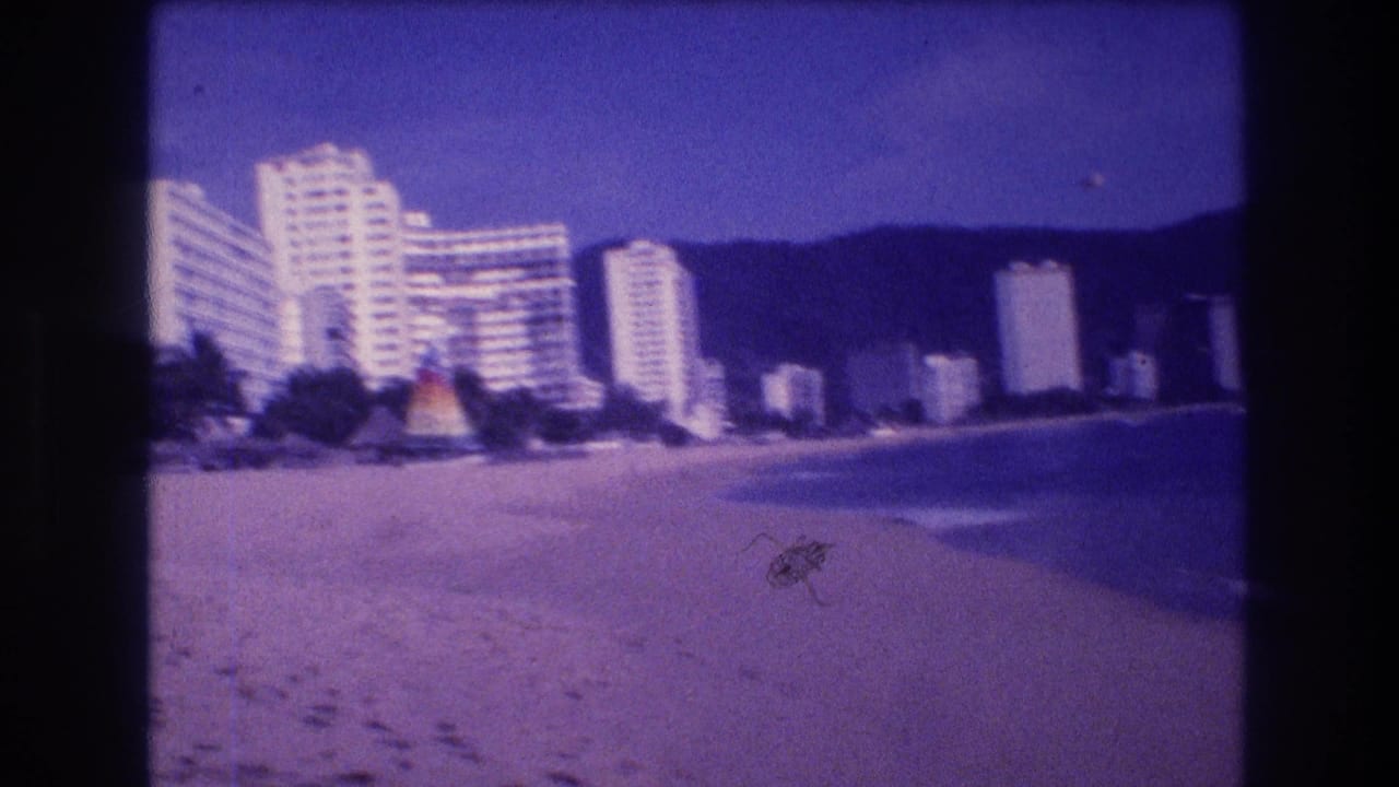 8mm home movie frame from beach level showing Acapulco hotel complex, colorful beach pavilion structure, palm trees, and footprints in sand, 1982.