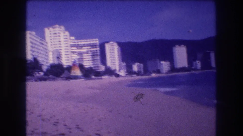 8mm home movie frame from beach level showing Acapulco hotel complex, colorful beach pavilion structure, palm trees, and footprints in sand, 1982.