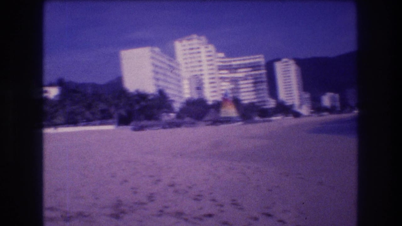 8mm home movie frame showing Acapulco hotel strip from the beach, colorful beach umbrella, palm trees, and footprints in foreground sand, 1982.