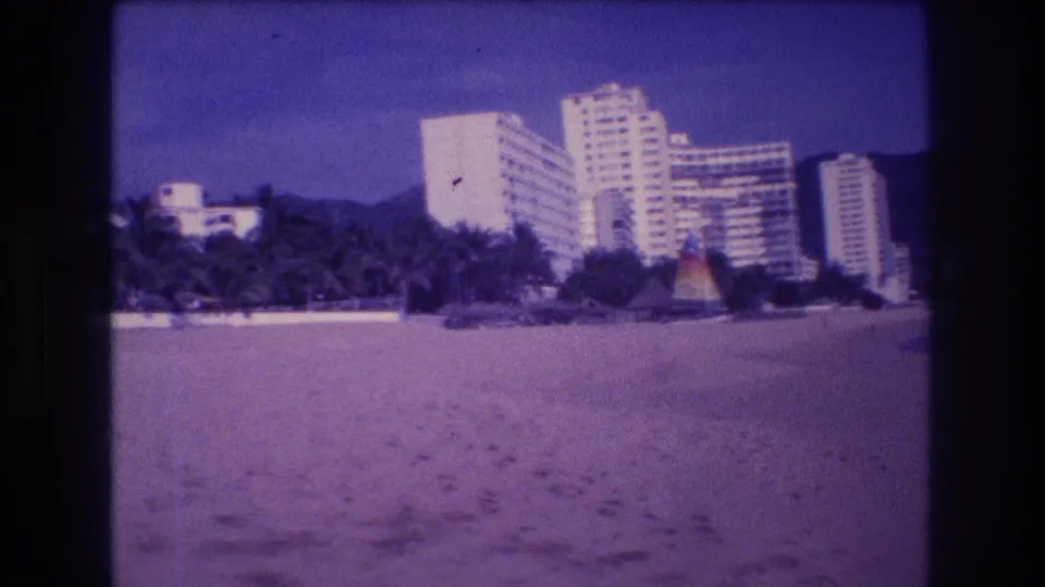Acapulco beachfront circa 1982: wide sandy beach, cluster of white resort high-rises, colorful palapas, and mountain backdrop on aged 8mm film with strong purple color shift.