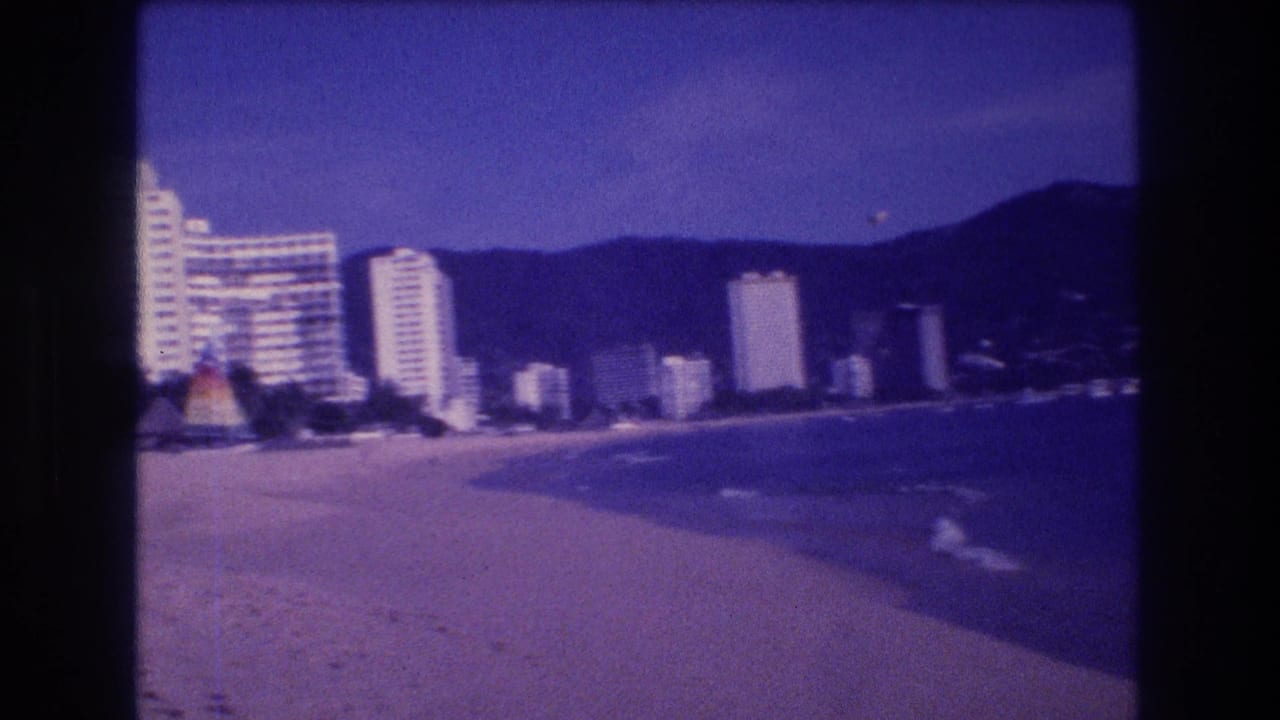 Panned view along Acapulco's beachfront showing white high-rise hotels, colorful palapas, curving coastline, and distant mountains; strong purple film cast.