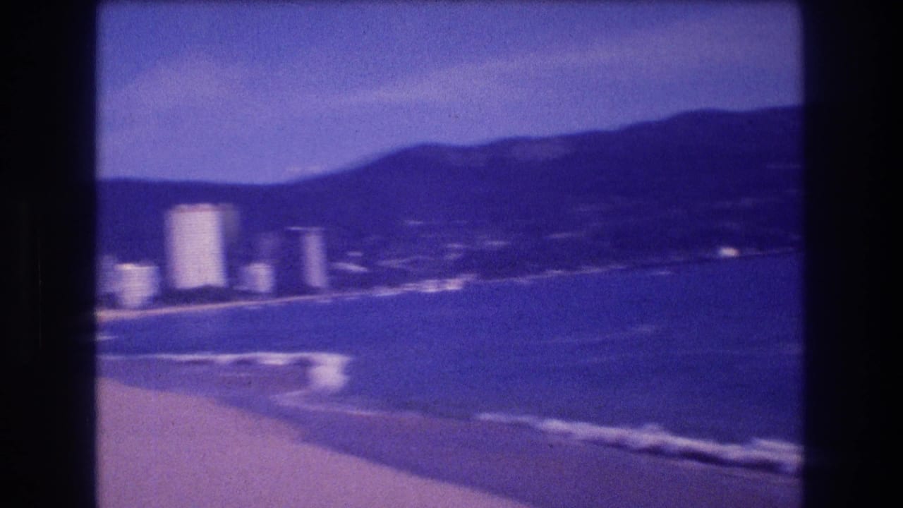 Acapulco bay panorama circa 1982: breaking waves on sandy shore, white buildings at the bay edge, mountain silhouette, and hazy sky on aged 8mm film with heavy purple color shift.