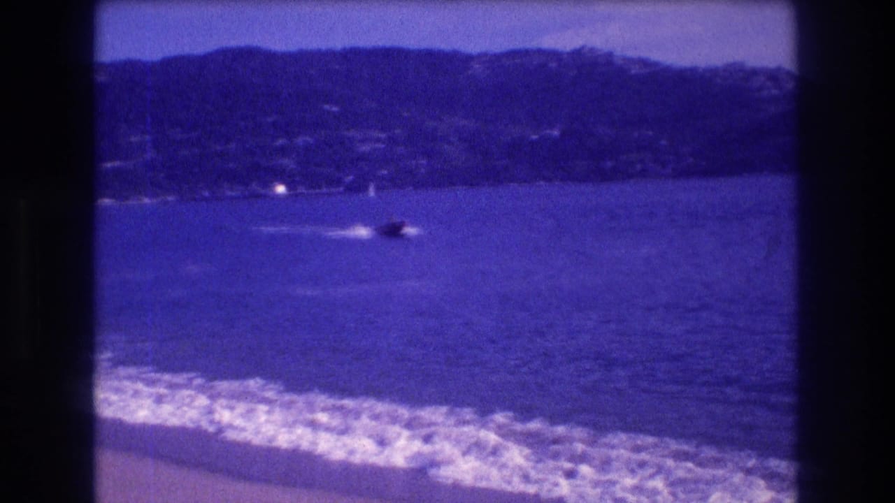 Acapulco shoreline circa 1982: waves wash the sand, a small boat crosses the bay, and a mountain range rises behind the water on visibly aged 8mm film.