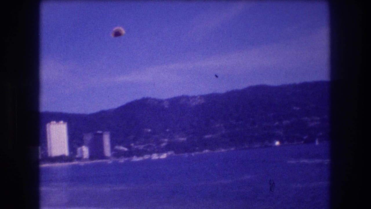 Parasail canopy airborne over coastal bay with high-rise hotel towers, sandy beach, and mountain ridge visible; aged blue 8mm film cast.