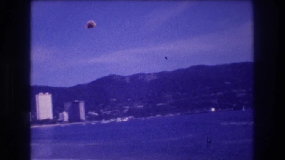 8mm home-movie frame circa 1982 showing a parasail ascending above Acapulco bay, with a strip of high-rise resort hotels, sandy beach, and mountains behind.