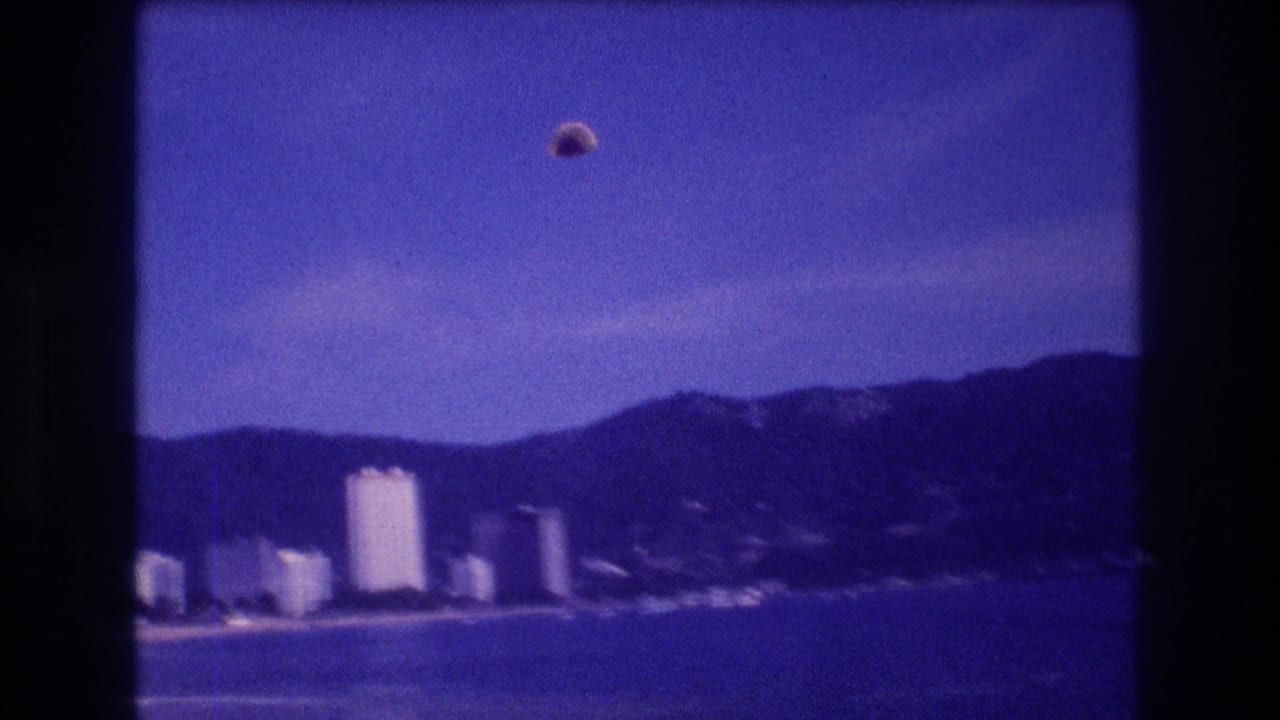 8mm home-movie frame circa 1982 of a parasail high above the Acapulco coastal hotel strip, with mountains and blue bay stretching below.