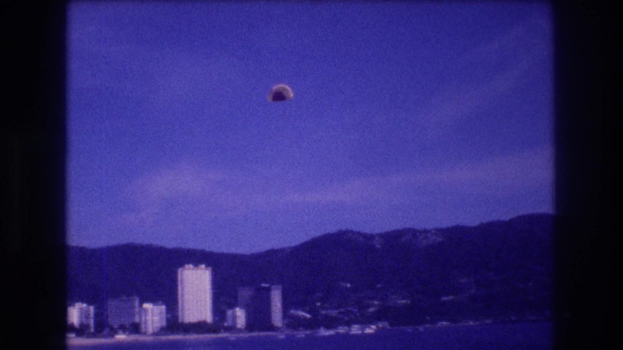 8mm home-movie frame circa 1982, parasail over Acapulco bay with the resort hotel skyline and mountain ridge visible in the distance.