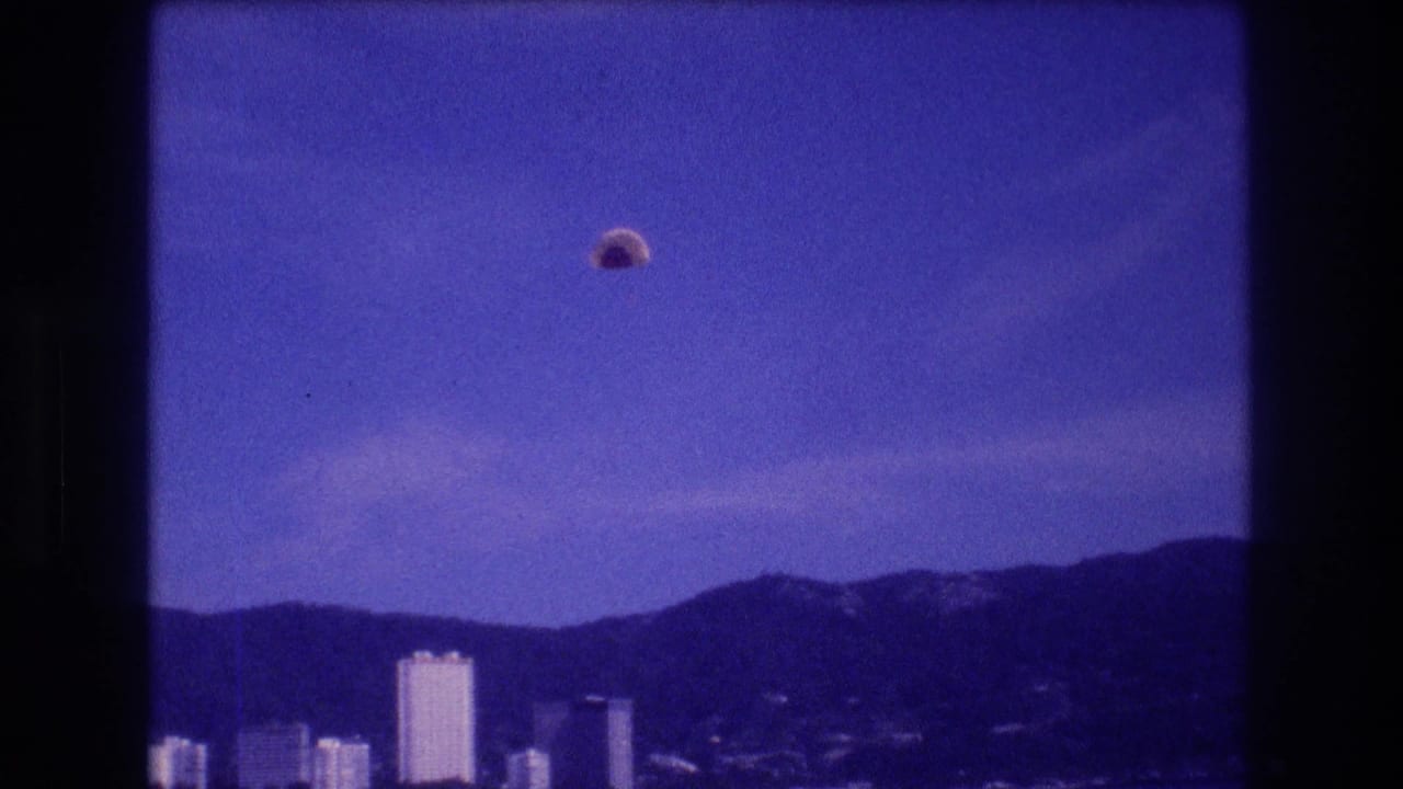 Blue-cast 8mm frame: small dome parachute canopy high above a coastal bay with white high-rise buildings and a mountain ridge in the background.