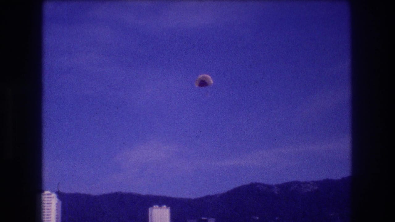 Blue-cast 8mm frame: dome parachute canopy slightly lower and to the right over the bay; white hotel towers and mountain ridge in background.