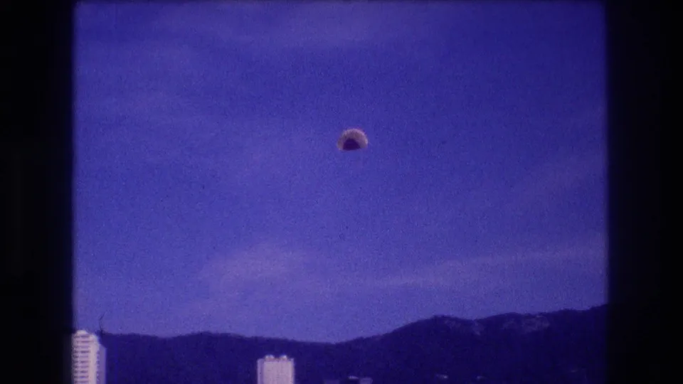 8mm home-movie frame, Acapulco Mexico c. 1982: parachute canopy mid-descent over the bay, hotel corridor and mountain backdrop visible.